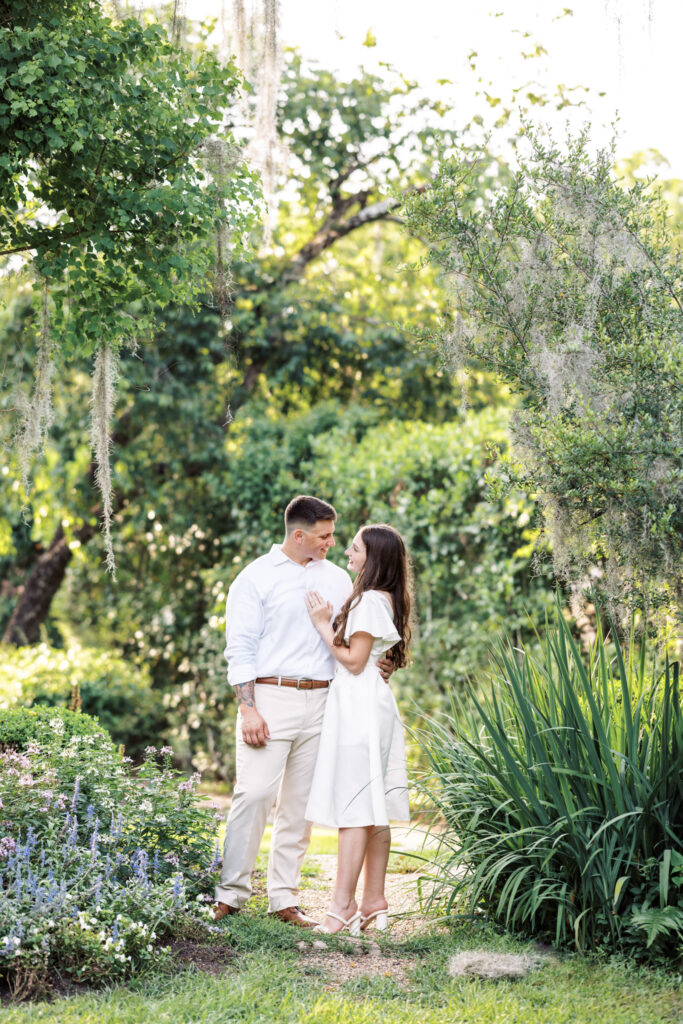 A couple dressed in white looking into each other's eyes in front of lush greenery at their Afton Villa Gardens engagement session