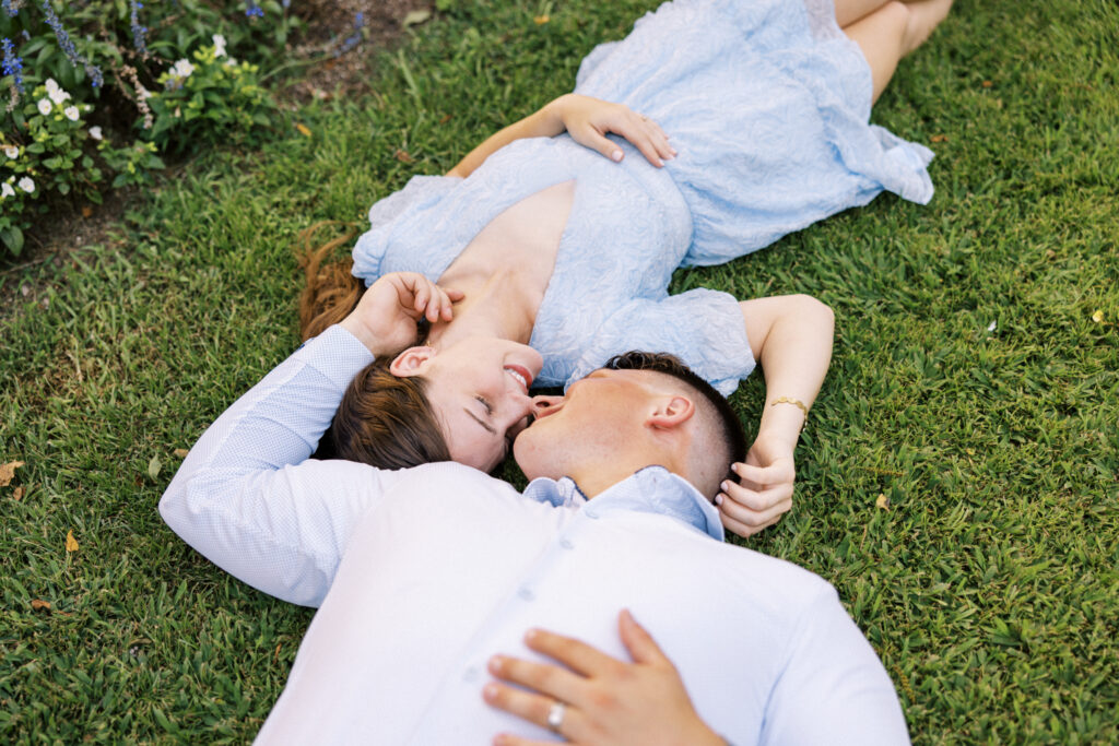 A couple dressed in soft blues laying on the grass in front of trees at their Afton Villa Gardens engagement session