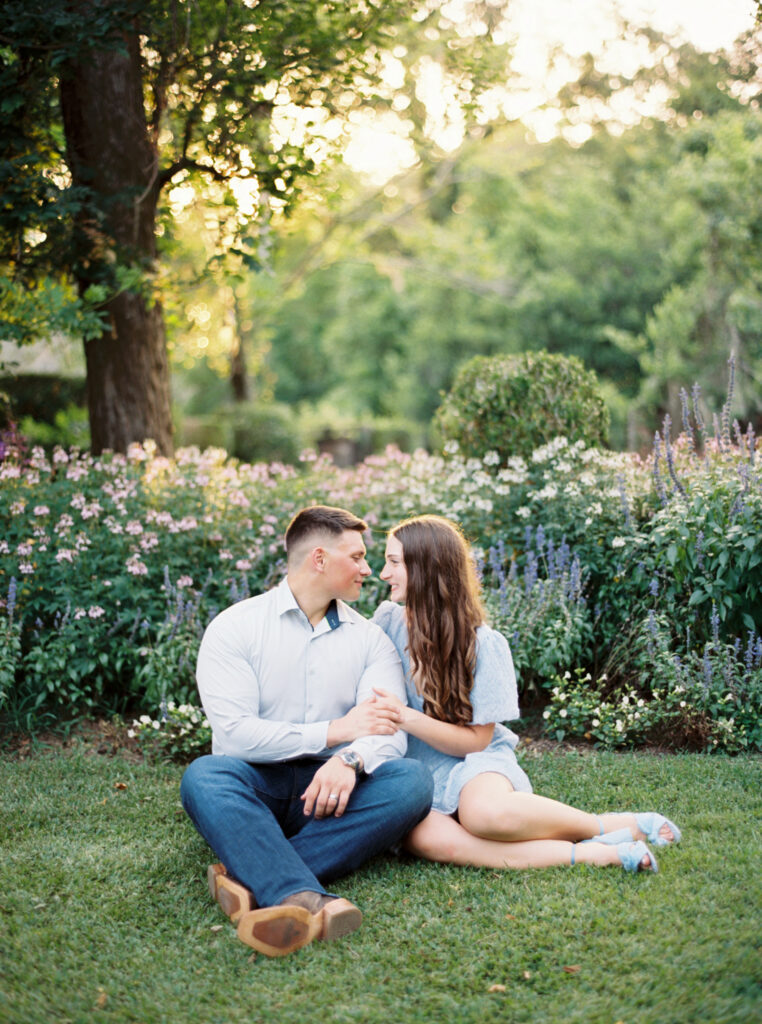 A couple dressed in soft blues sitting on the grass and looking into each other's eyes at their Afton Villa Gardens engagement session