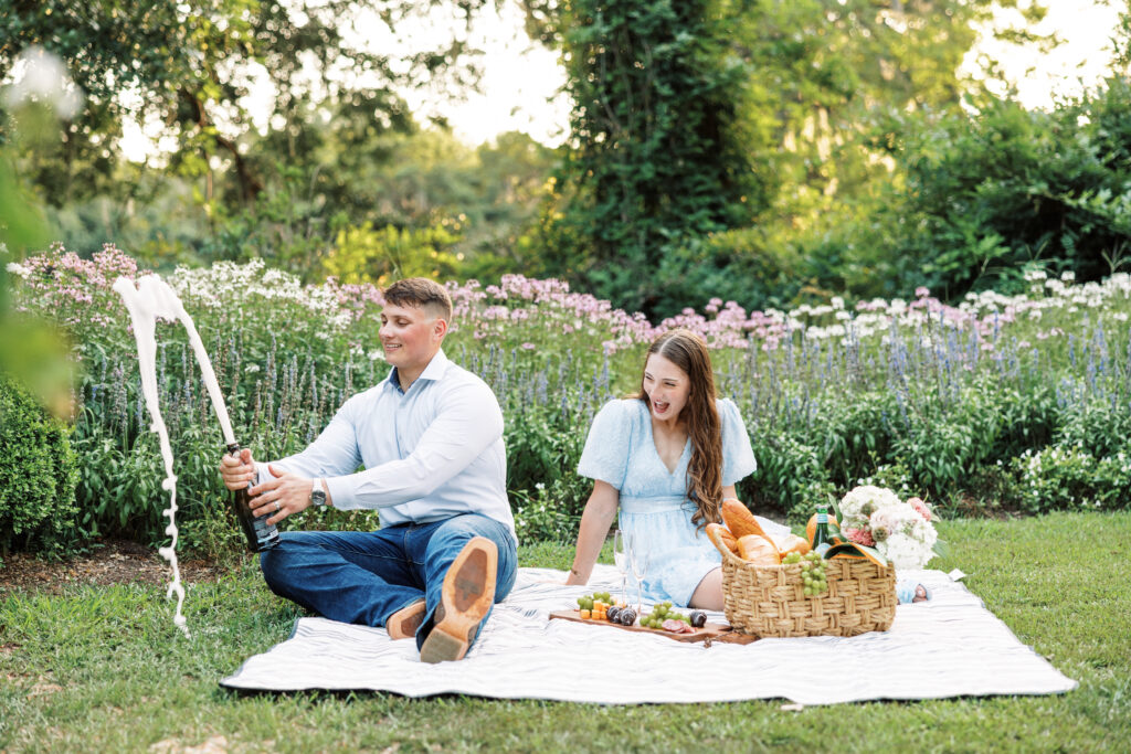 A couple dressed in soft blues having a picnic on the grass with champagne at their Afton Villa Gardens engagement session