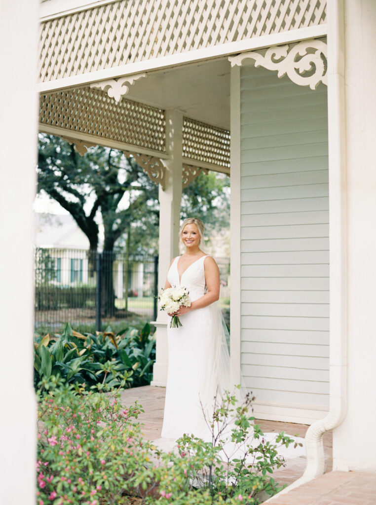 A bridal portrait at John Nickerson House in Lafayette, LA, showing a smiling bride in a white gown and veil holding a bouquet while standing on a Victorian balcony.