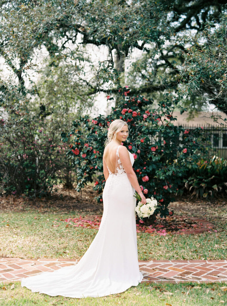 A bridal portrait at John Nickerson House in Lafayette, LA, showing a smiling bride in a white gown holding a bouquet while standing in front of lush greenery and pink flowers.