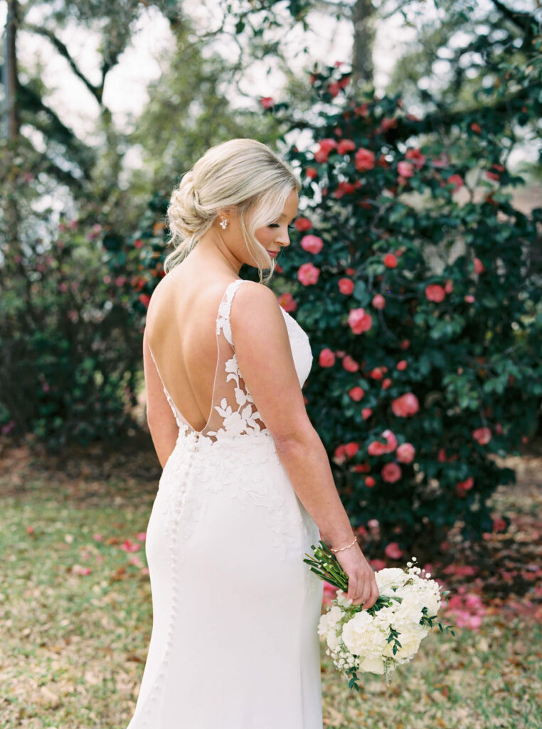 A bridal portrait at John Nickerson House in Lafayette, LA, showing a bride in a white gown holding a bouquet while standing in front of lush greenery and pink flowers.