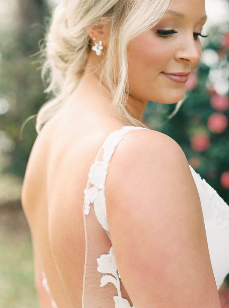 A bridal portrait at John Nickerson House in Lafayette, LA, showing a smiling bride in a white gown and veil standing in front of lush greenery and pink flowers.