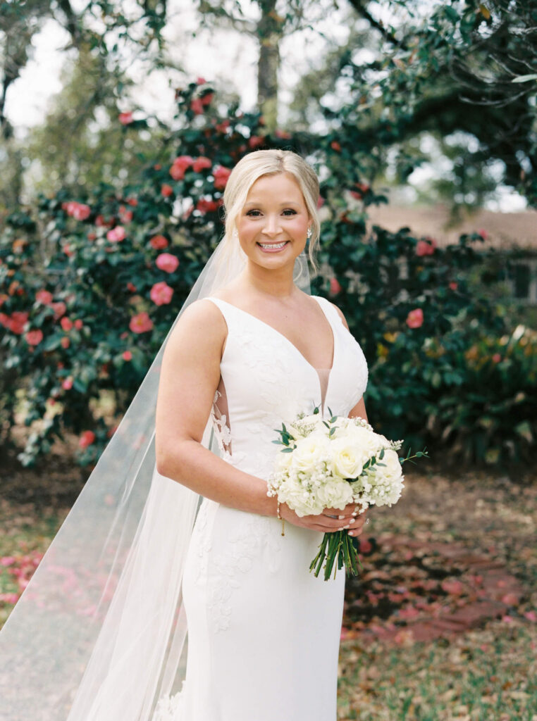 A bridal portrait at John Nickerson House in Lafayette, LA, showing a smiling bride in a white gown and veil holding a bouquet while standing in front of lush greenery and pink flowers.