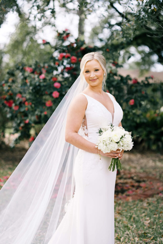 A bridal portrait at John Nickerson House in Lafayette, LA, showing a smiling bride in a white gown and veil holding a bouquet while standing in front of lush greenery and pink flowers.