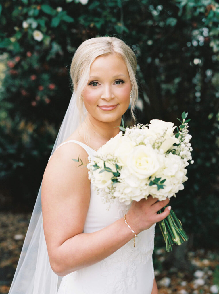 A bridal portrait at John Nickerson House in Lafayette, LA, showing a smiling bride in a white gown and veil holding a bouquet while standing in front of lush greenery.