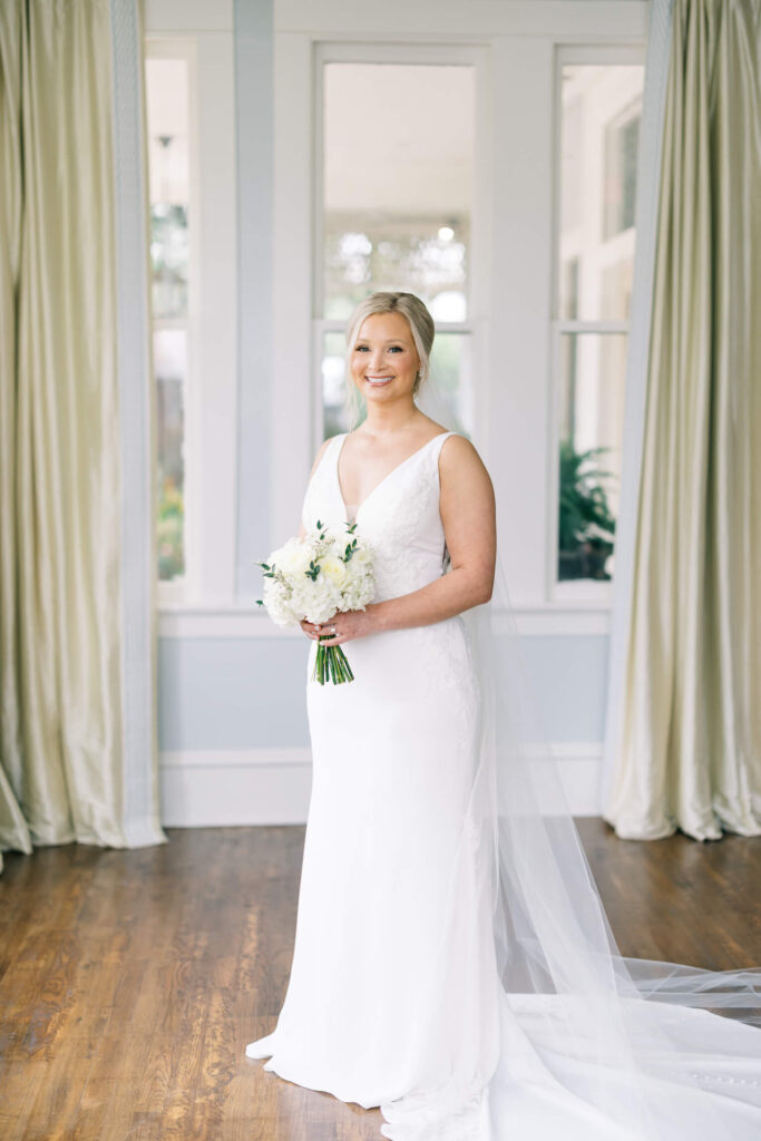 A bridal portrait at John Nickerson House in Lafayette, LA, showing a smiling bride in a white gown and veil holding a bouquet while standing in front of a tall window.