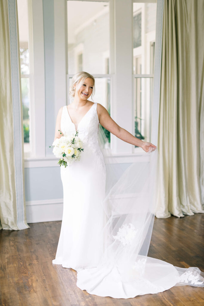 A bridal portrait at John Nickerson House in Lafayette, LA, showing a smiling bride in a white gown and veil holding a bouquet while standing in front of a tall window.