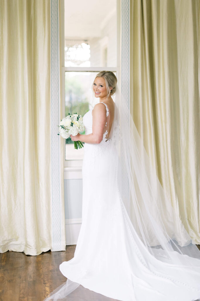 A bridal portrait at John Nickerson House in Lafayette, LA, showing a smiling bride in a white gown and veil holding a bouquet while standing in front of a tall window.