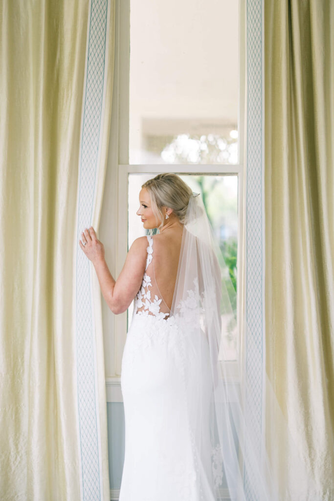 A bridal portrait at John Nickerson House in Lafayette, LA, showing a smiling bride in a white gown and veil looking out a tall window.