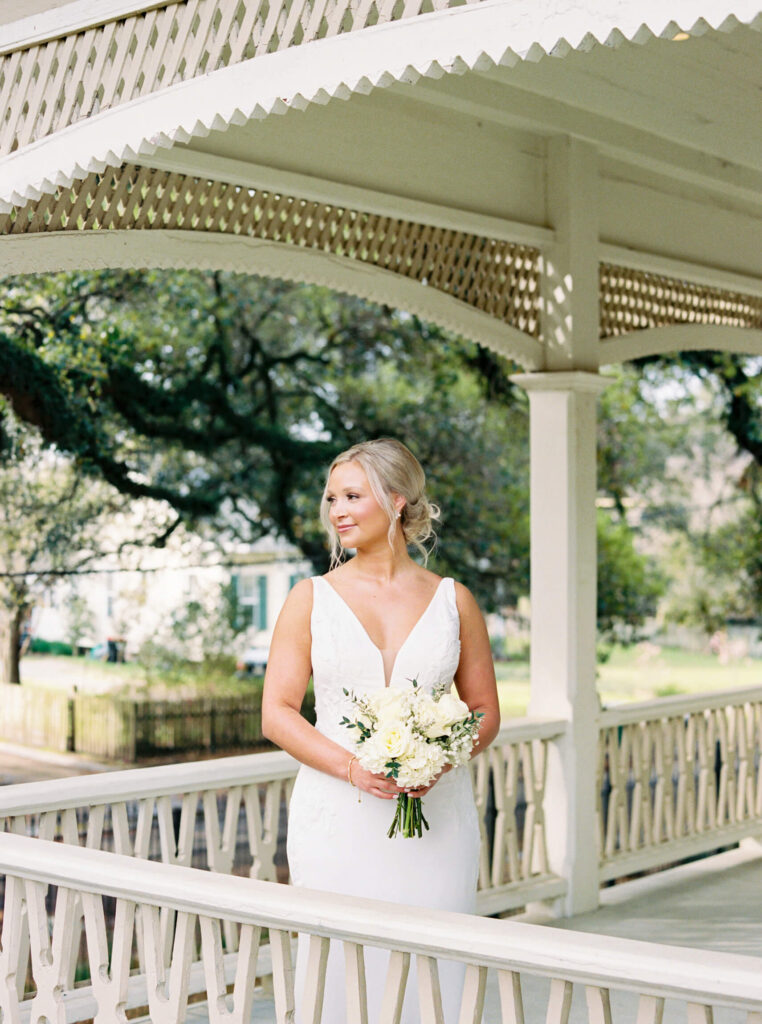 A bridal portrait at John Nickerson House in Lafayette, LA, showing a smiling bride in a white gown holding a bouquet while overlooking the porch.