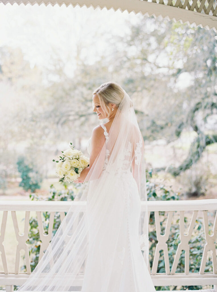 A bridal portrait at John Nickerson House in Lafayette, LA, showing a smiling bride in a white gown and veil holding a bouquet while overlooking the porch.