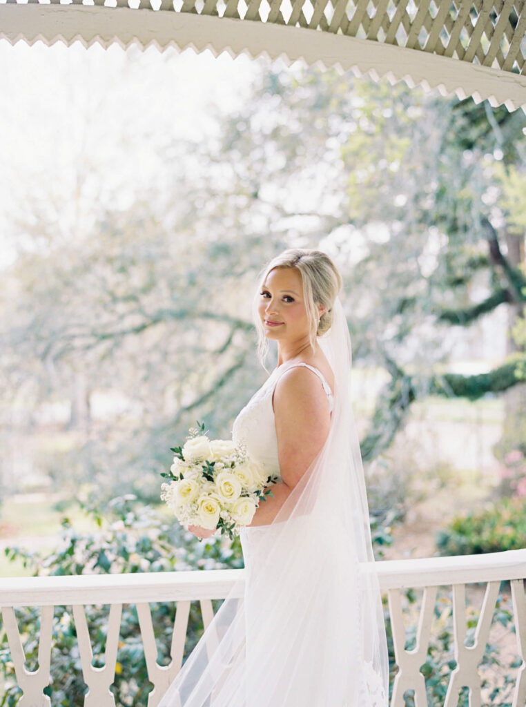 A bridal portrait at John Nickerson House in Lafayette, LA, showing a smiling bride in a white gown and veil holding a bouquet while overlooking the porch.