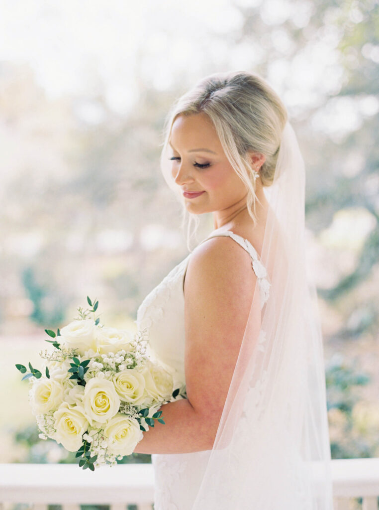 A bridal portrait at John Nickerson House in Lafayette, LA, showing a smiling bride in a white gown and veil holding a bouquet while overlooking the porch.