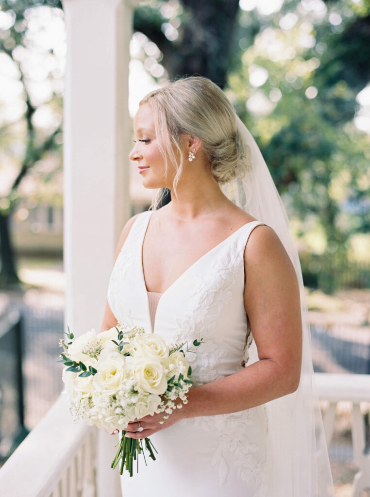 A bridal portrait at John Nickerson House in Lafayette, LA, showing a smiling bride in a white gown and veil holding a bouquet while overlooking the porch.