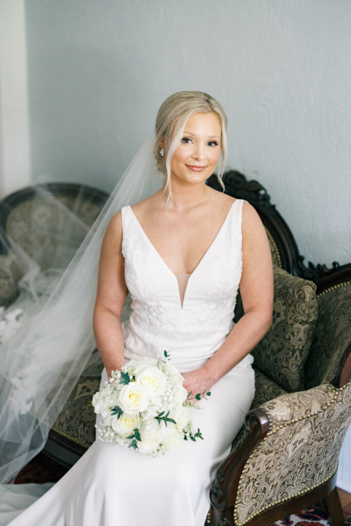 A bridal portrait at John Nickerson House in Lafayette, LA, showing a smiling bride in a white gown and veil holding a bouquet while sitting on a Victorian couch.