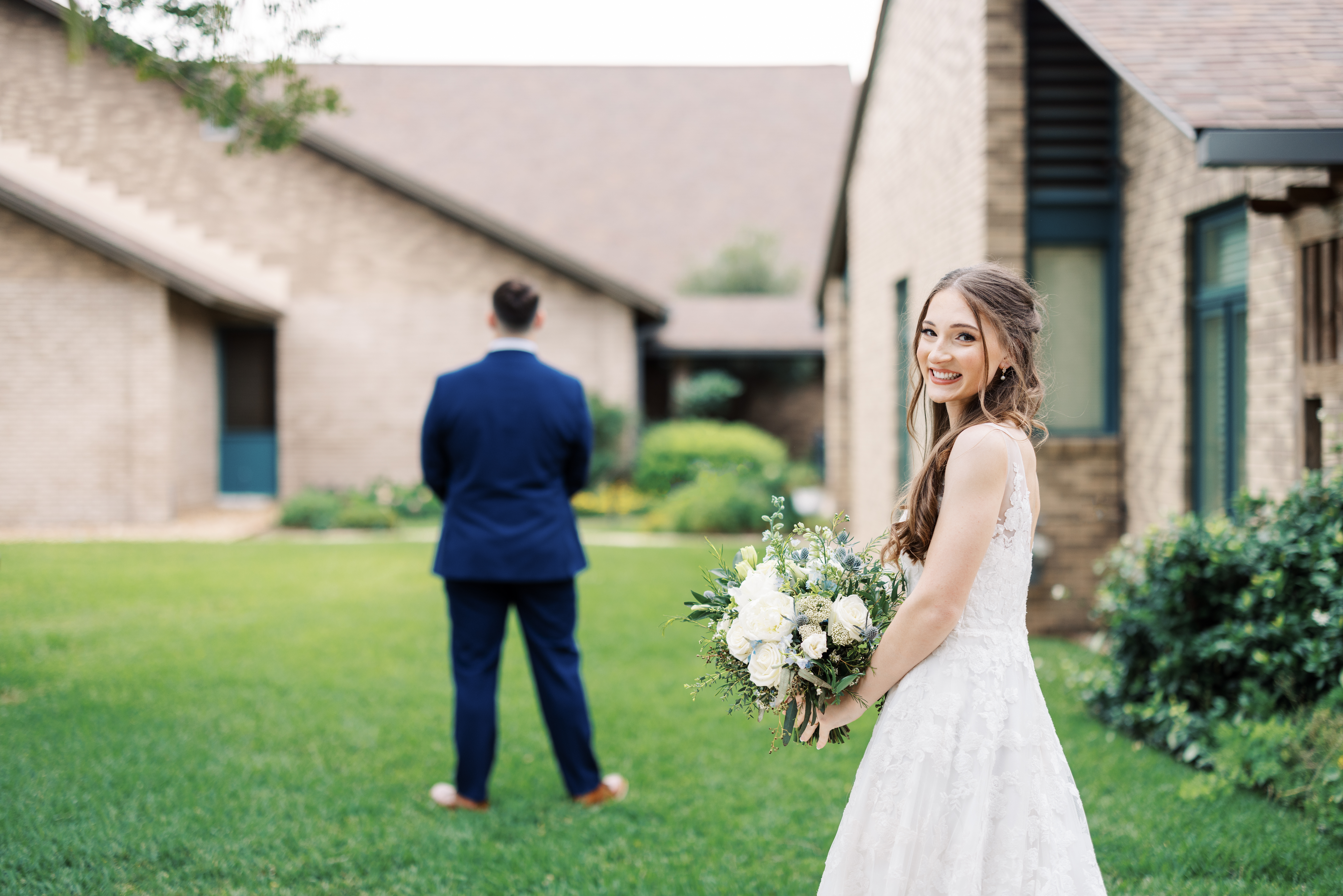 A bride in white holding a bouquet smiling towards the camera while the groom in a blue suit is turned away, before a wedding day first look