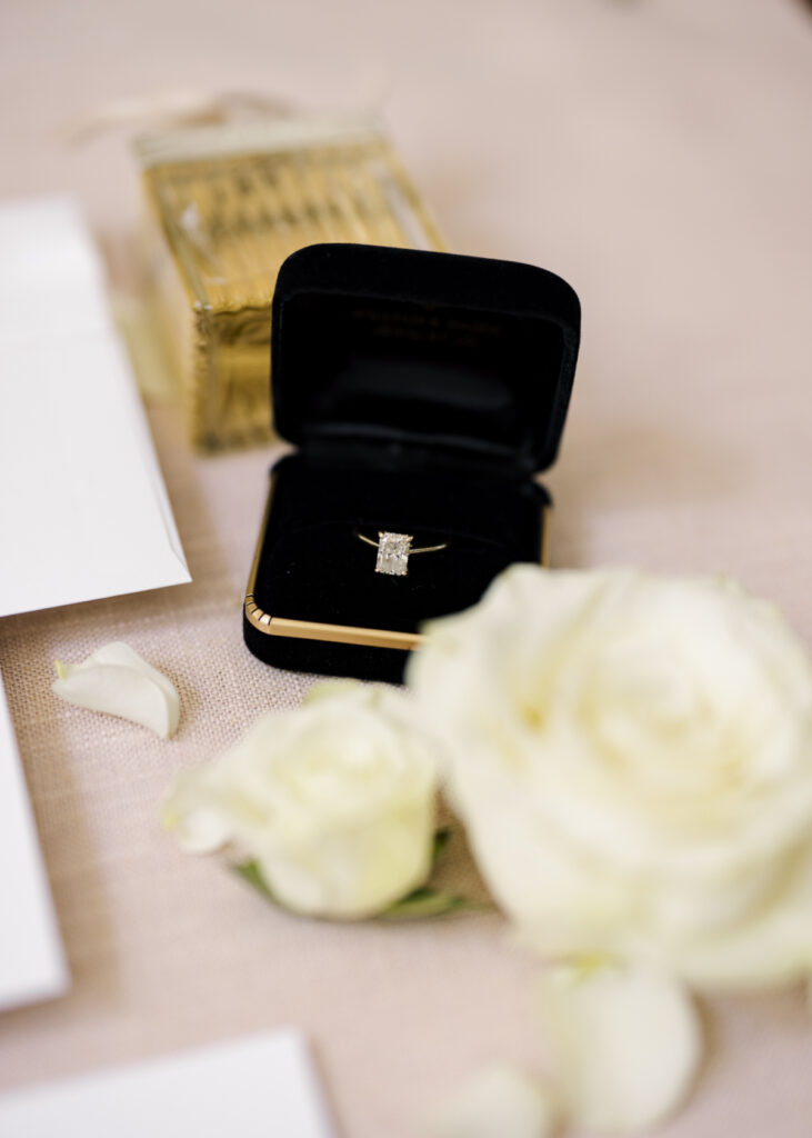 Radiant cut diamond engagement ring in a black and gold ring box with white flowers in a wedding details shot by Morgan Alysse Photography