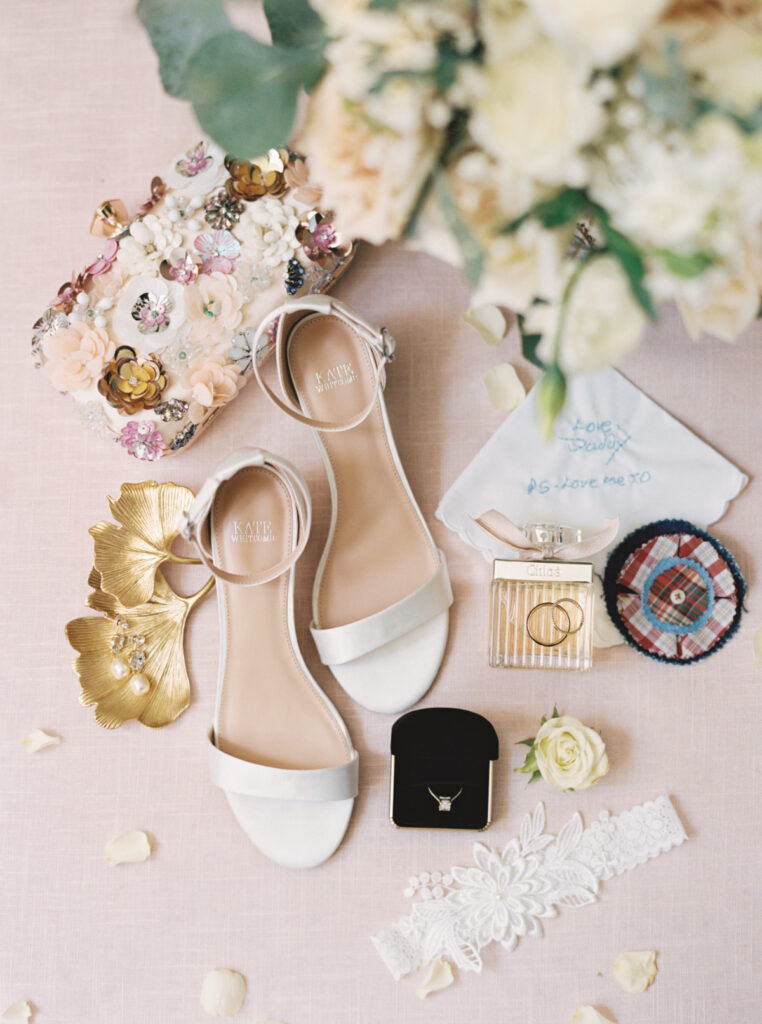 Wedding shoes, a floral beaded clutch, perfume, and a black and gold ring box with white flowers in a wedding details shot by Morgan Alysse Photography